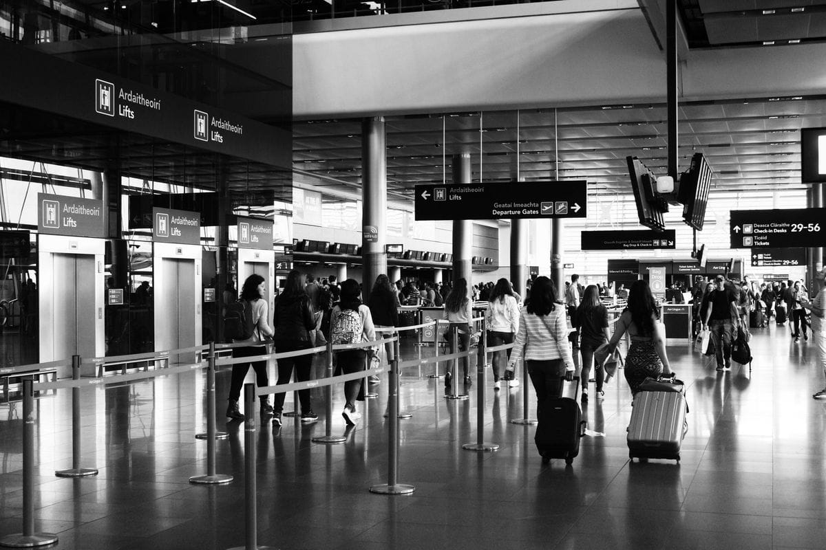 Black and white photo of travellers pulling cabin suitcases past queue barriers in an airport terminal