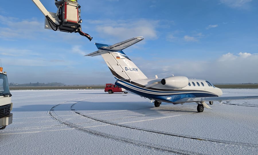 De-icing truck boom raised beside a small jet on a snowy, frosted airport apron under a blue sky