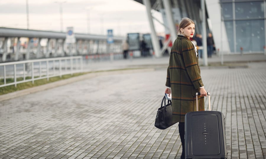 Traveller in a long checked wool coat pulling a dark suitcase outside an airport terminal