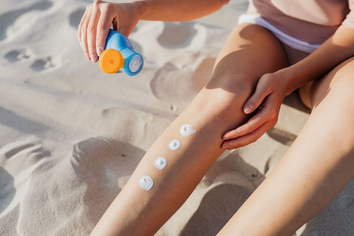 Person sitting on a sandy beach squeezing dots of sunscreen onto their leg from a blue bottle