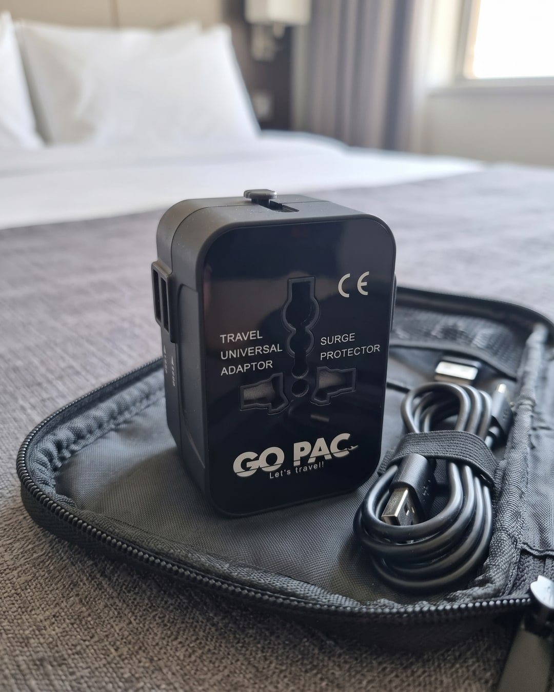 Black GO PAC travel adapter resting on an open zip pouch holding coiled charging cables, on a hotel bed