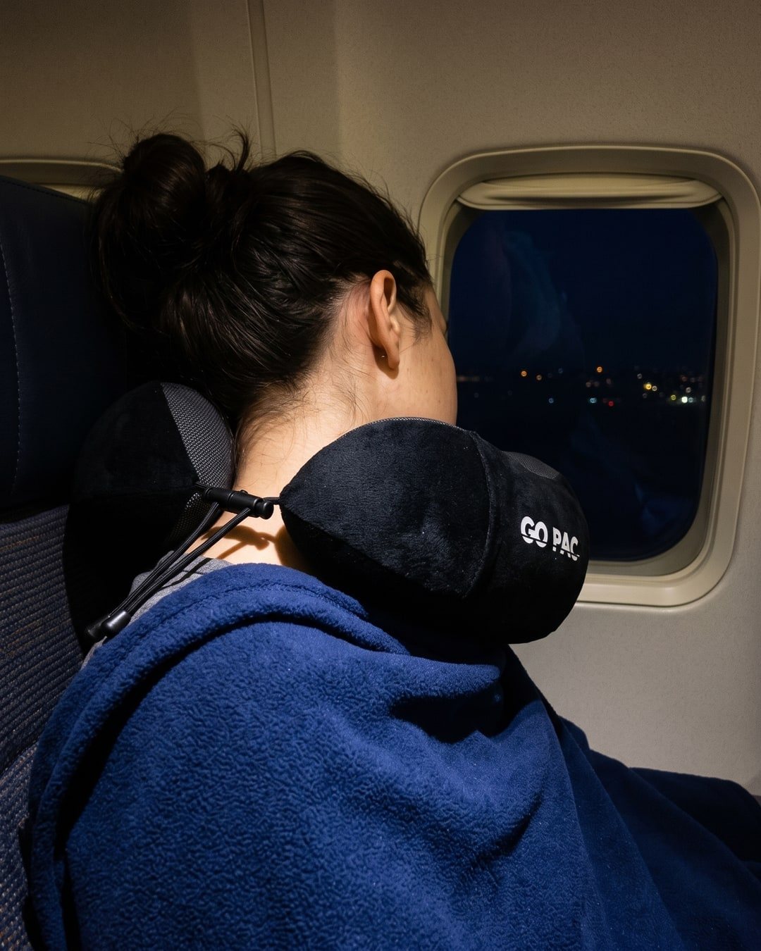 Traveller asleep against a plane window at night wearing a black GO PAC memory foam travel pillow