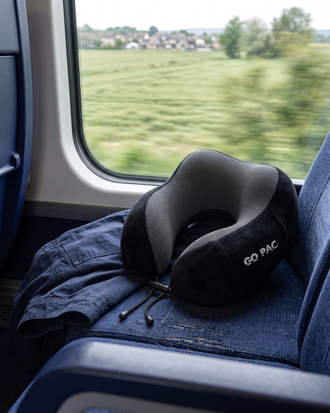 Black GO PAC memory foam travel pillow on a train seat by the window with fields passing outside